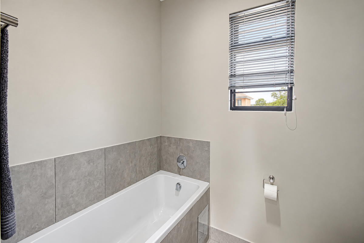 A clean and modern bathroom featuring a built-in bathtub with grey tiled surround, chrome fixtures, and a window with venetian blinds.