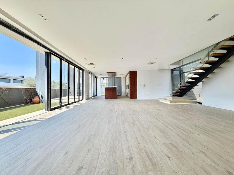 Spacious open-plan living area featuring modern laminate flooring, a sleek staircase with glass balustrades, and large sliding doors opening to the garden.
