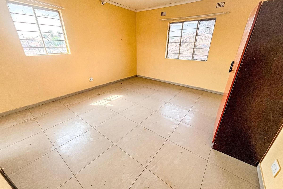 An empty bedroom featuring tiled flooring, large windows with burglar bars, and yellow painted walls, offering a blank canvas for renovation.