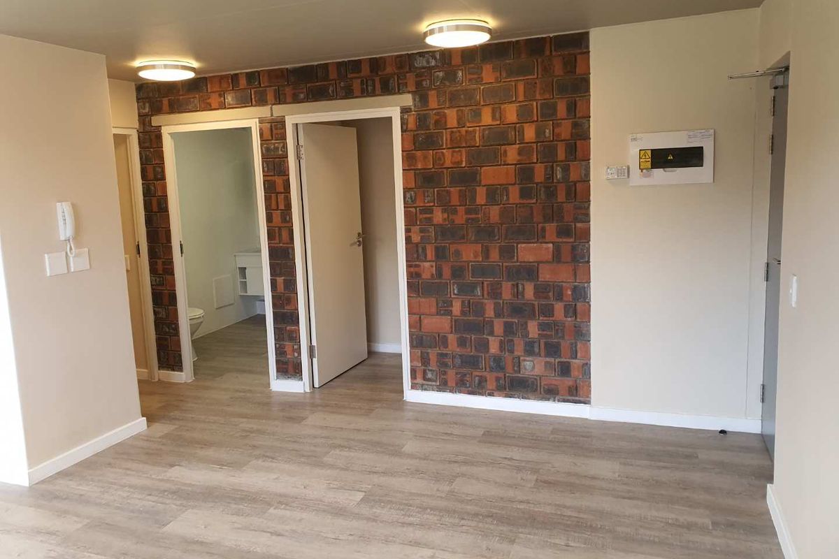 An open-plan living area featuring a distinctive exposed brick wall, modern laminate flooring, and neutral finishes, providing a versatile space for customization.