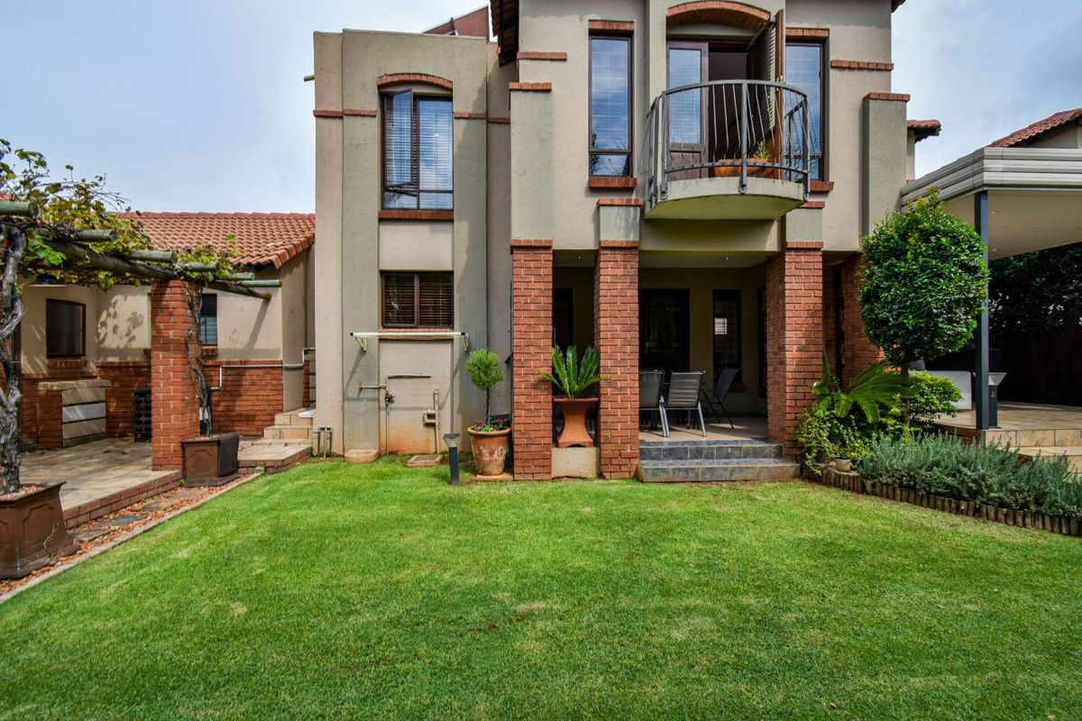Rear view of the townhouse featuring a manicured lawn, covered patio area, brick pillar detailing, and an upper-level balcony with wrought iron railing.