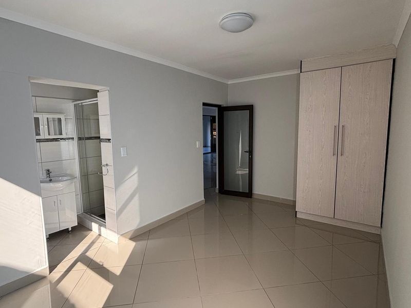 A clean, empty bedroom featuring tiled flooring, a built-in wardrobe, and an attached en-suite bathroom with a shower and vanity.
