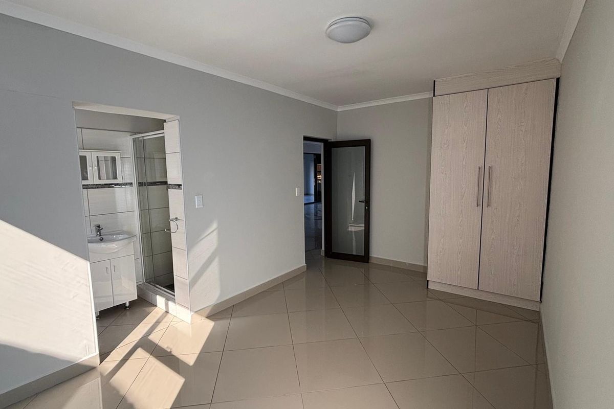 A clean, empty bedroom featuring tiled flooring, a built-in wardrobe, and an attached en-suite bathroom with a shower and vanity.
