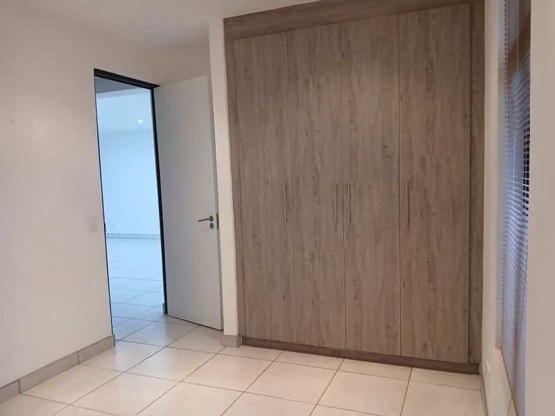 A clean, empty bedroom featuring modern wood-finish built-in cupboards, neutral tiled flooring, and venetian blinds, offering a bright and versatile living space.