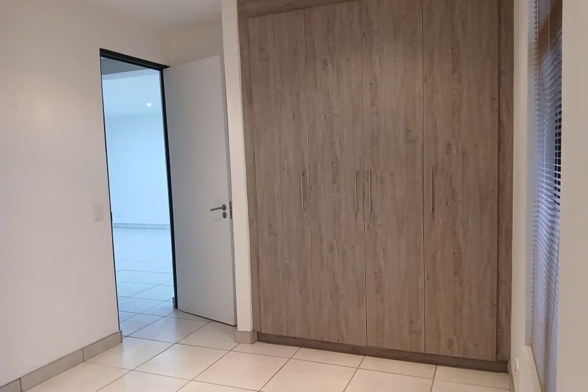 A clean, empty bedroom featuring modern wood-finish built-in cupboards, neutral tiled flooring, and venetian blinds, offering a bright and versatile living space.