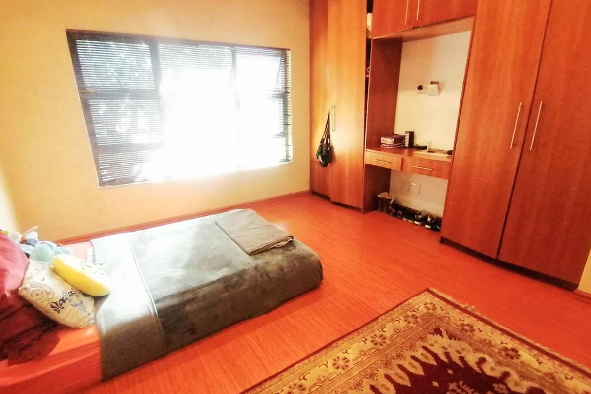 A bedroom featuring warm-toned laminate flooring, extensive built-in wooden cabinetry with a central vanity area, and large windows fitted with venetian blinds.