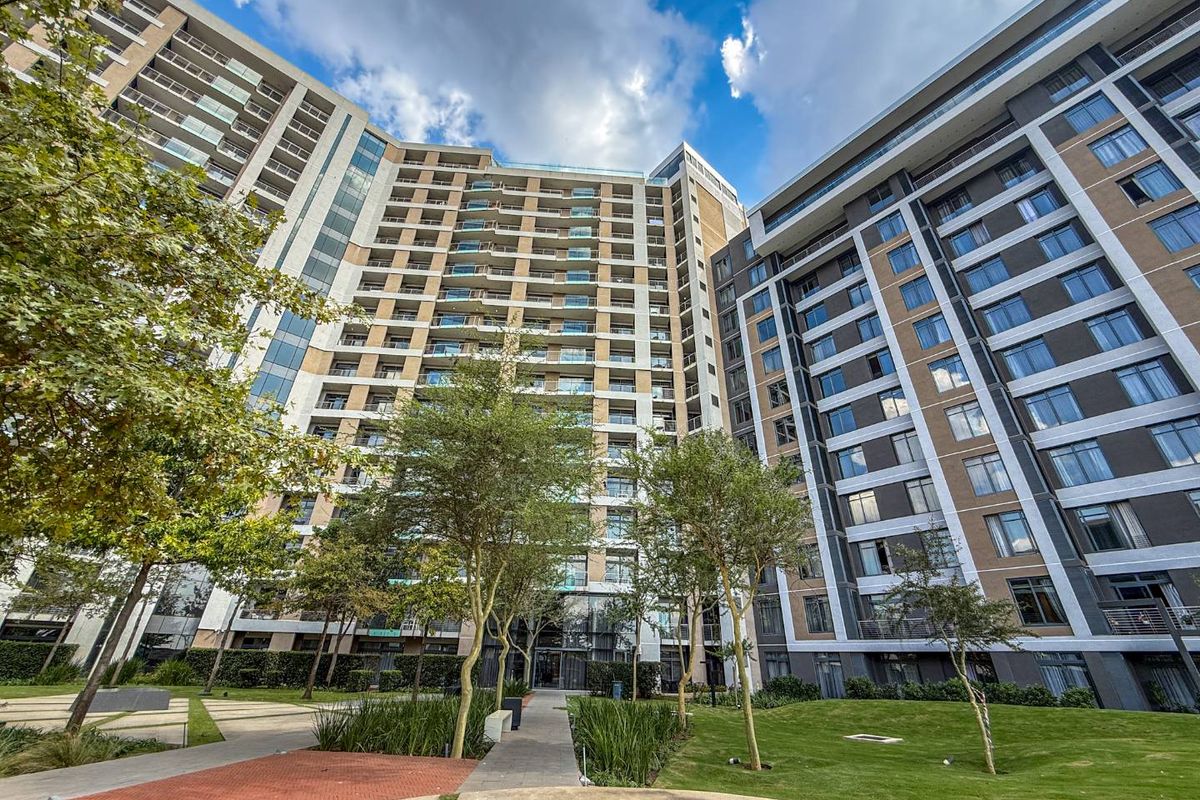 A modern high-rise apartment complex featuring a landscaped garden, paved walkways, and multiple levels of private balconies.