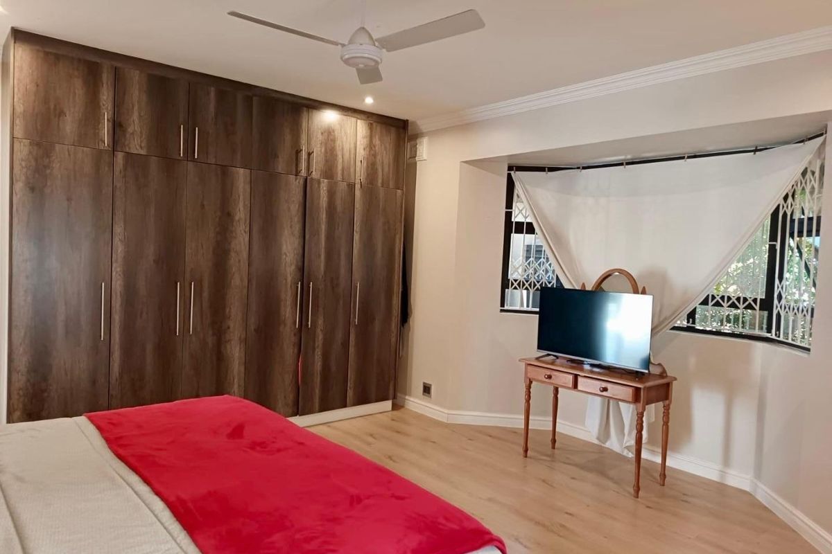 Spacious bedroom featuring extensive built-in wooden cupboards, light laminate flooring, a ceiling fan, and a bay window with security gate.