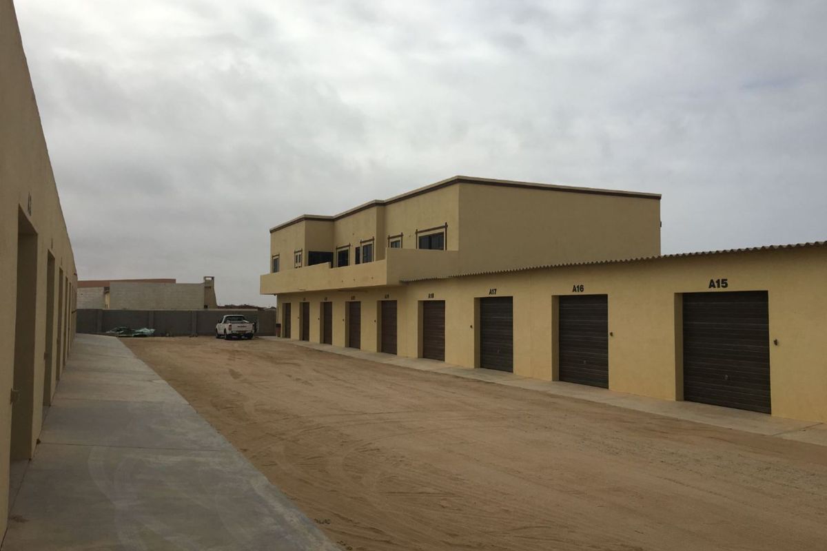 Exterior view of an industrial complex featuring a row of numbered garage units with a sandy driveway and a multi-level building structure.