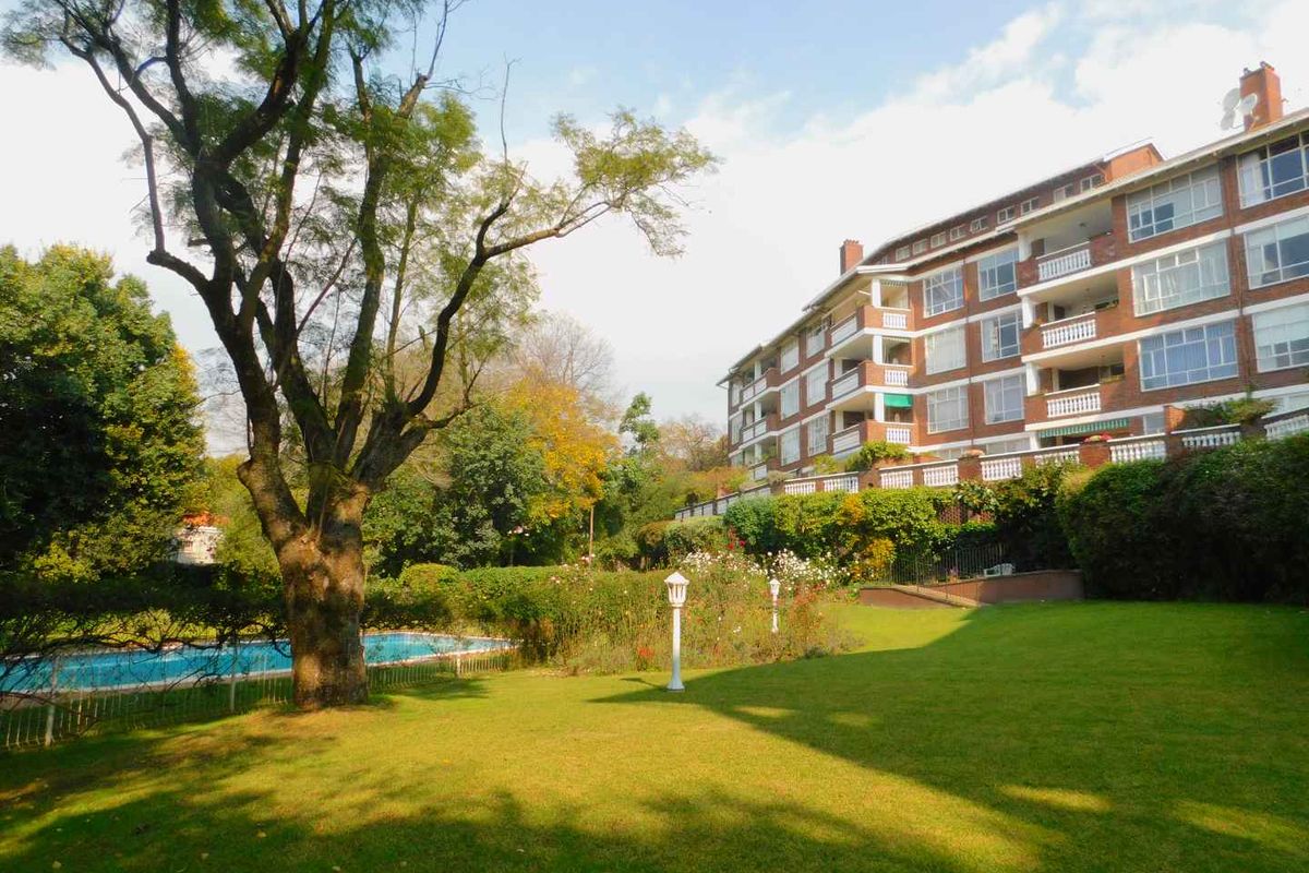 A view of the apartment complex rear facade featuring a well-maintained garden, swimming pool, and established trees.