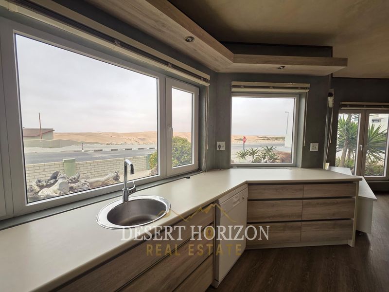 Modern kitchen area featuring light wood-finish cabinetry, laminate countertops, a stainless steel sink, and large windows offering scenic desert views.