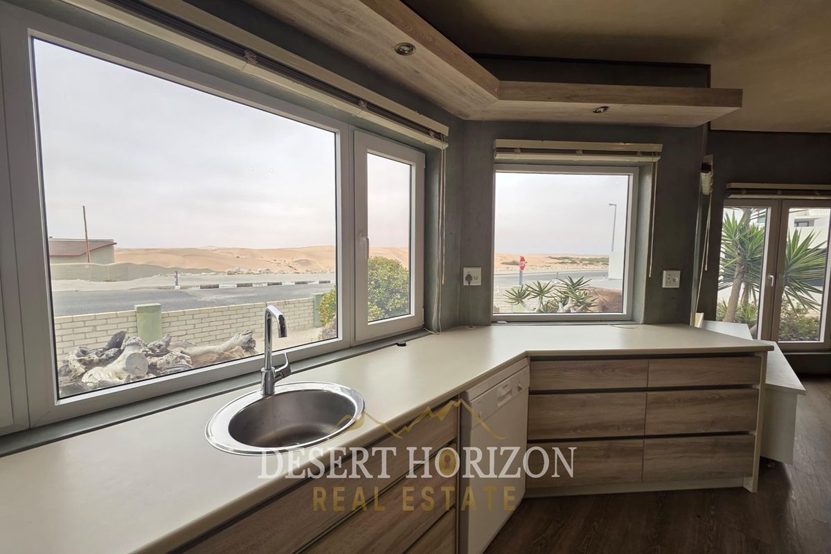 Modern kitchen area featuring light wood-finish cabinetry, laminate countertops, a stainless steel sink, and large windows offering scenic desert views.