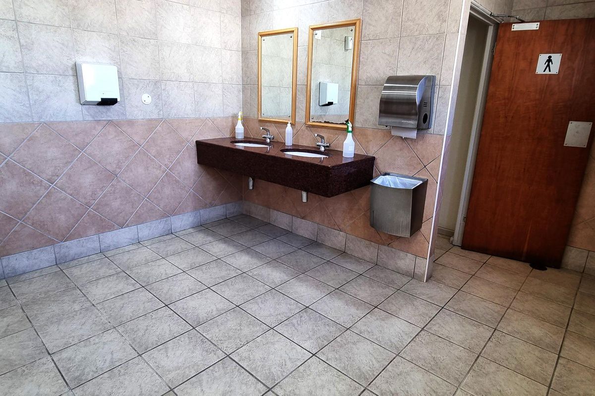 A functional office restroom featuring a double vanity with granite countertop, wall-mounted mirrors, and stainless steel soap and paper towel dispensers.