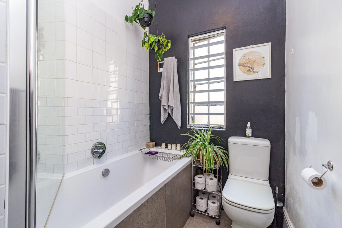 A compact bathroom featuring a bathtub with a shower screen, a modern toilet, and a louver window with contrasting dark and white wall finishes.