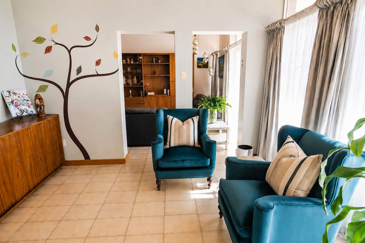 A bright living area featuring tiled flooring, large windows with curtains, and an open-plan layout leading into an adjacent room with built-in shelving.