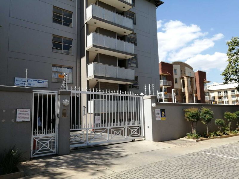 Front entrance view of Llonon Crest apartment complex featuring a secure gated access point, perimeter wall with electric fencing, and multi-story residential balconies.