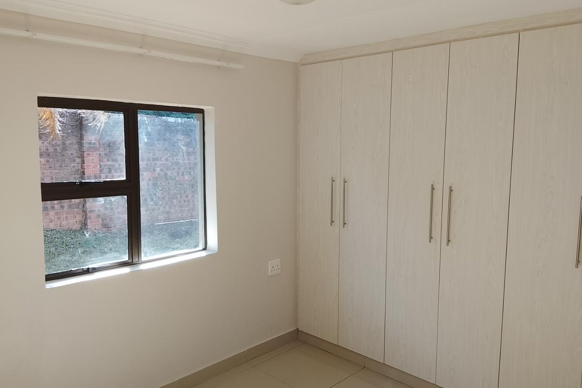 A clean, empty bedroom featuring neutral tiled flooring, built-in light wood-grain cupboards, and an aluminium-framed window overlooking the exterior.