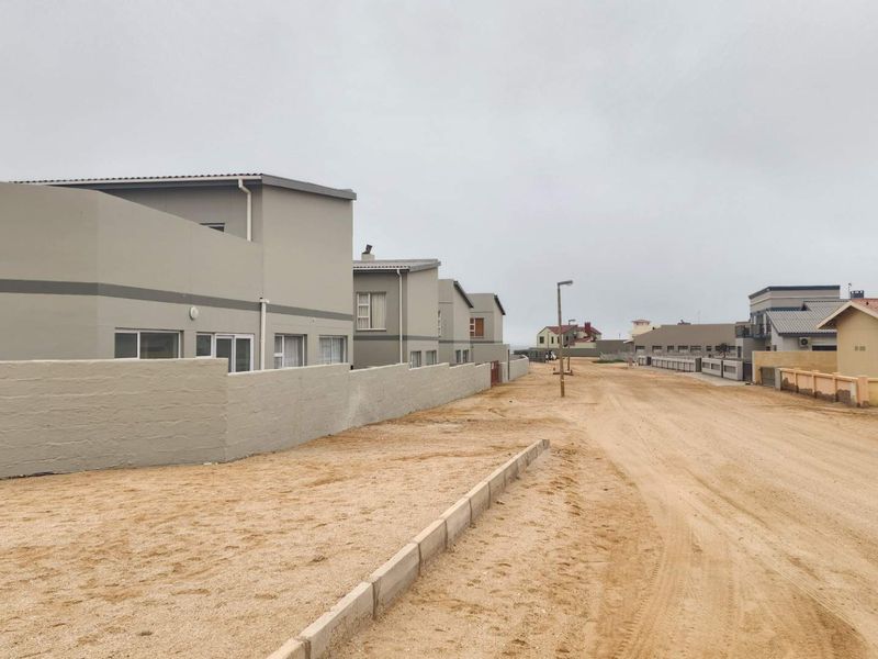 A street view of a residential townhouse development featuring modern grey facades, boundary walls, and an unpaved sandy road surface.