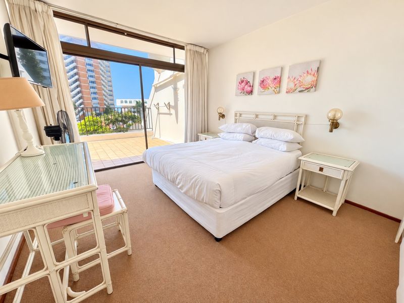A bright bedroom featuring carpeted flooring, wall-mounted lighting, and a large sliding glass door providing direct access to a private balcony.