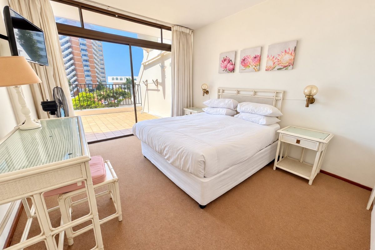 A bright bedroom featuring carpeted flooring, wall-mounted lighting, and a large sliding glass door providing direct access to a private balcony.