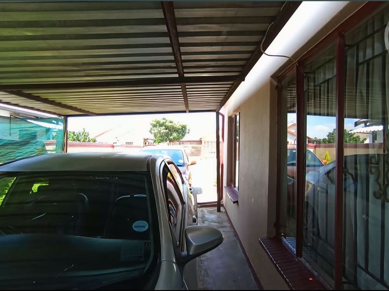 A covered carport or patio area featuring a corrugated metal roof and large windows with security bars along the side of the house.