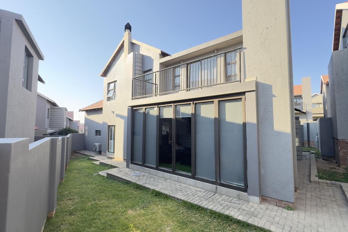 Rear view of a modern two-story house featuring a paved patio, a balcony with metal railings, and large glass sliding doors.