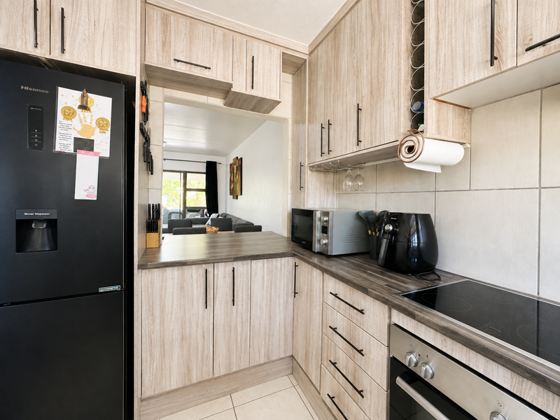A well-maintained kitchen featuring light wood-grain cabinetry, dark laminate countertops, a built-in oven, electric hob, and a convenient pass-through window to the living area.