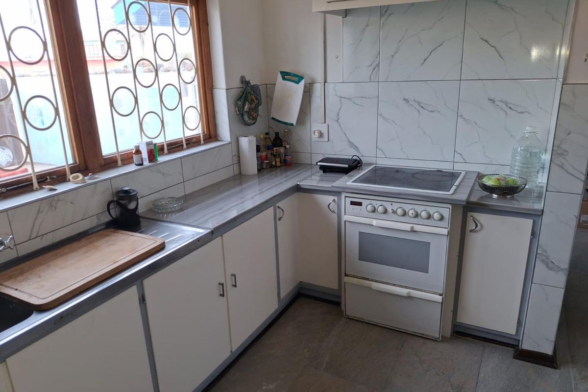 A functional kitchen featuring white cabinetry, marble-effect wall tiles, a built-in oven with hob, and a window with security bars.