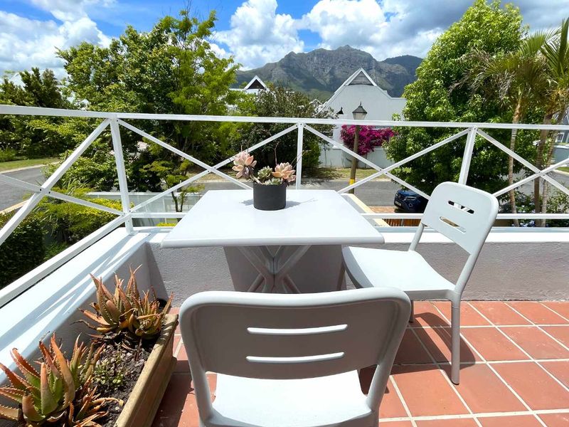 A sunny balcony featuring terracotta tiling and a white metal balustrade, offering pleasant views of the surrounding neighborhood and mountain backdrop.