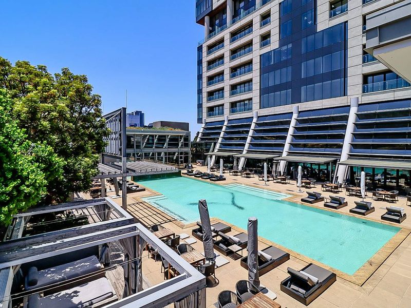 A well-maintained outdoor swimming pool area featuring a large pool, timber pergolas, and paved decking, set against a modern high-rise apartment building.