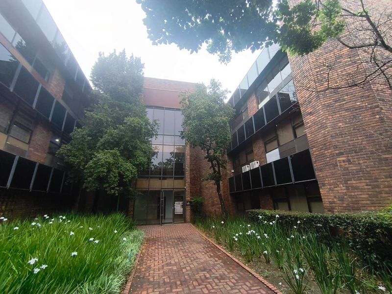 A multi-story office building featuring a brick facade, glass entrance atrium, and a paved walkway leading through a landscaped garden area.