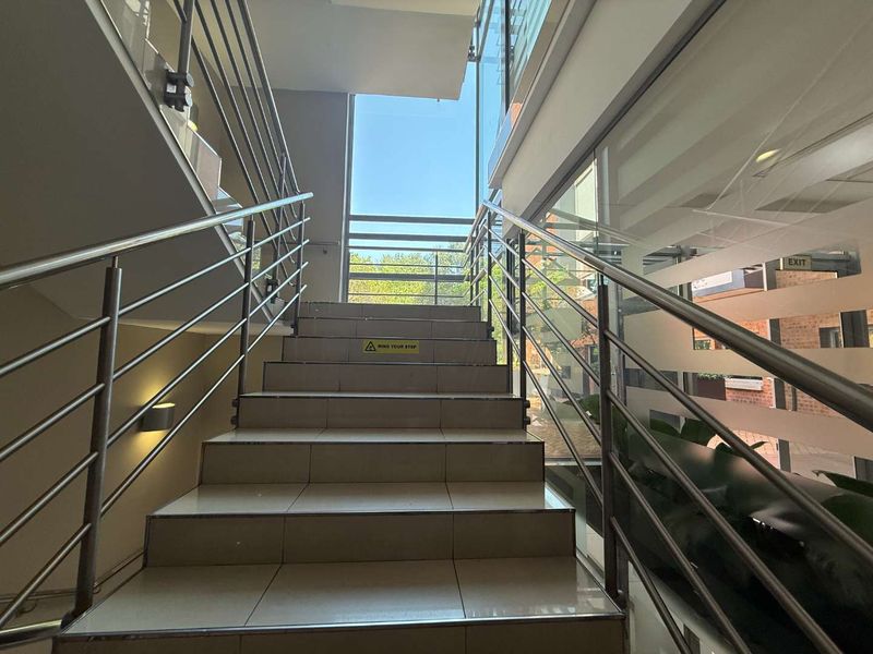 A bright, modern office staircase featuring tiled steps, stainless steel handrails, and a large glass window providing natural light.
