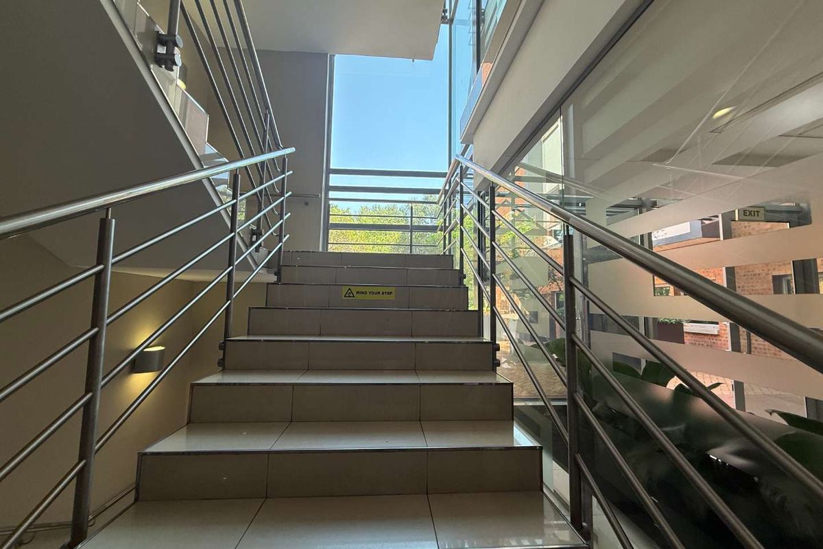 A bright, modern office staircase featuring tiled steps, stainless steel handrails, and a large glass window providing natural light.
