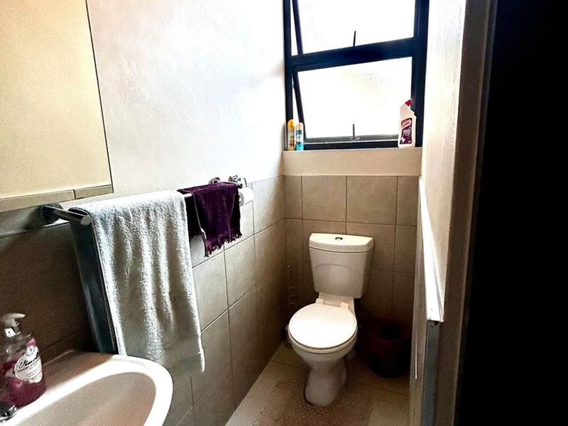 A compact bathroom featuring a pedestal basin, toilet, and neutral wall and floor tiling with a top-hung window for natural light.