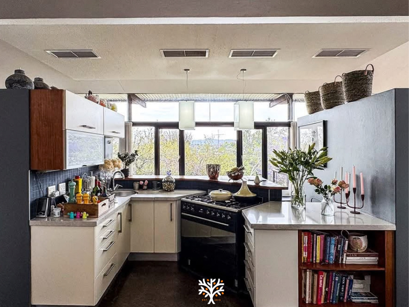 A compact, modern kitchen featuring built-in cabinetry, stone countertops, a gas stove, and large windows offering natural light and scenic views.