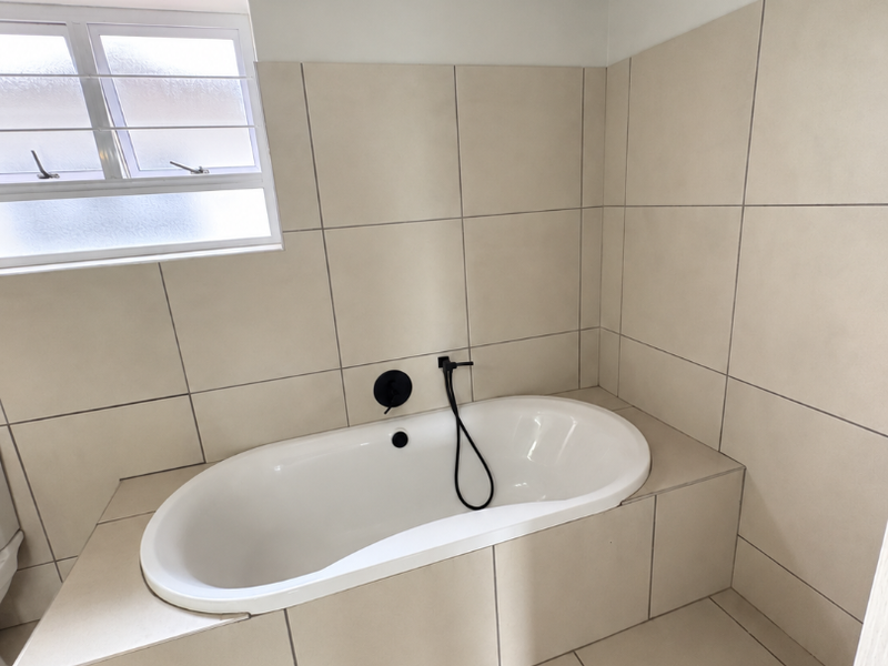 A modern bathroom featuring a built-in bathtub with matte black fixtures, neutral floor-to-ceiling tiling, and exposed timber ceiling beams.