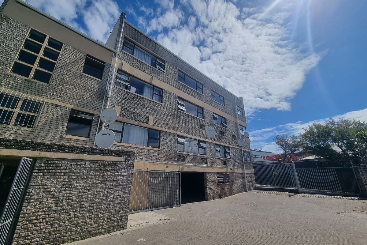 Exterior view of a multi-story apartment building featuring a brick facade, paved parking area, and secure metal gate access.