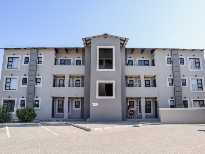 Front elevation of a modern multi-story apartment complex featuring paved parking, neutral facade finishes, and aluminum-framed windows.