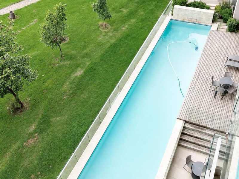 An elevated view of a modern rectangular swimming pool featuring a glass balustrade, timber decking, and stone paving, set within a well-maintained garden area.