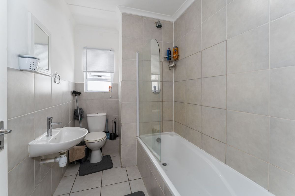 A functional bathroom featuring a wall-mounted basin, toilet, and a bathtub with a glass shower screen, finished with neutral wall and floor tiles.