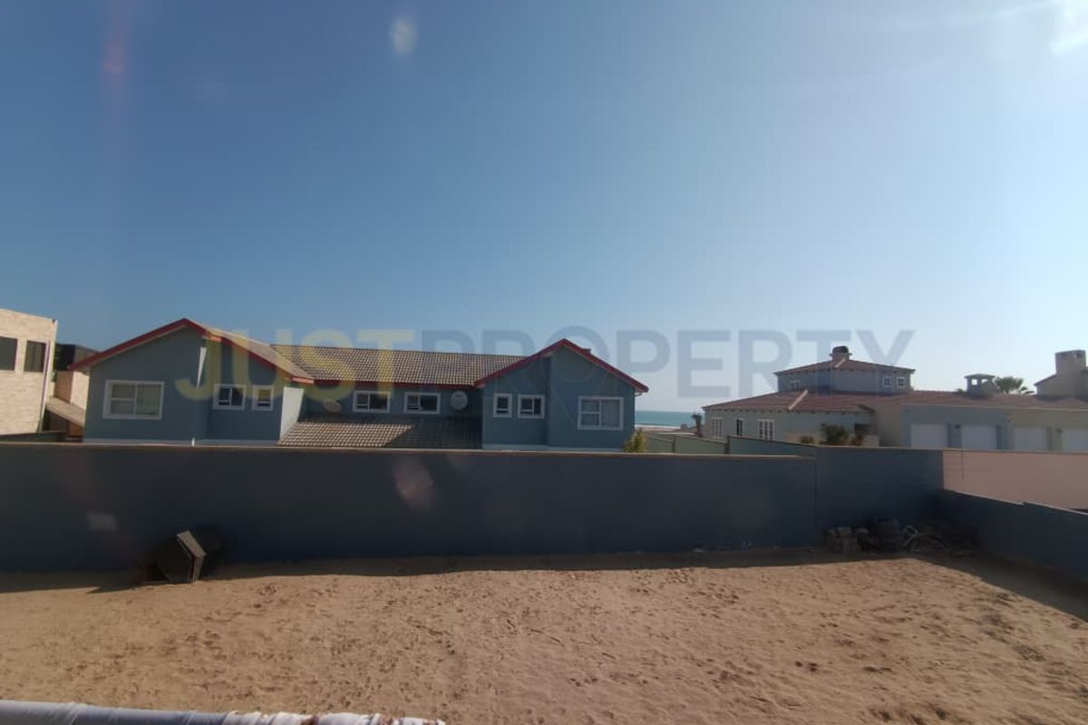 Rear view of the property showing a sandy backyard, boundary wall, and neighboring residential buildings under a clear sky.
