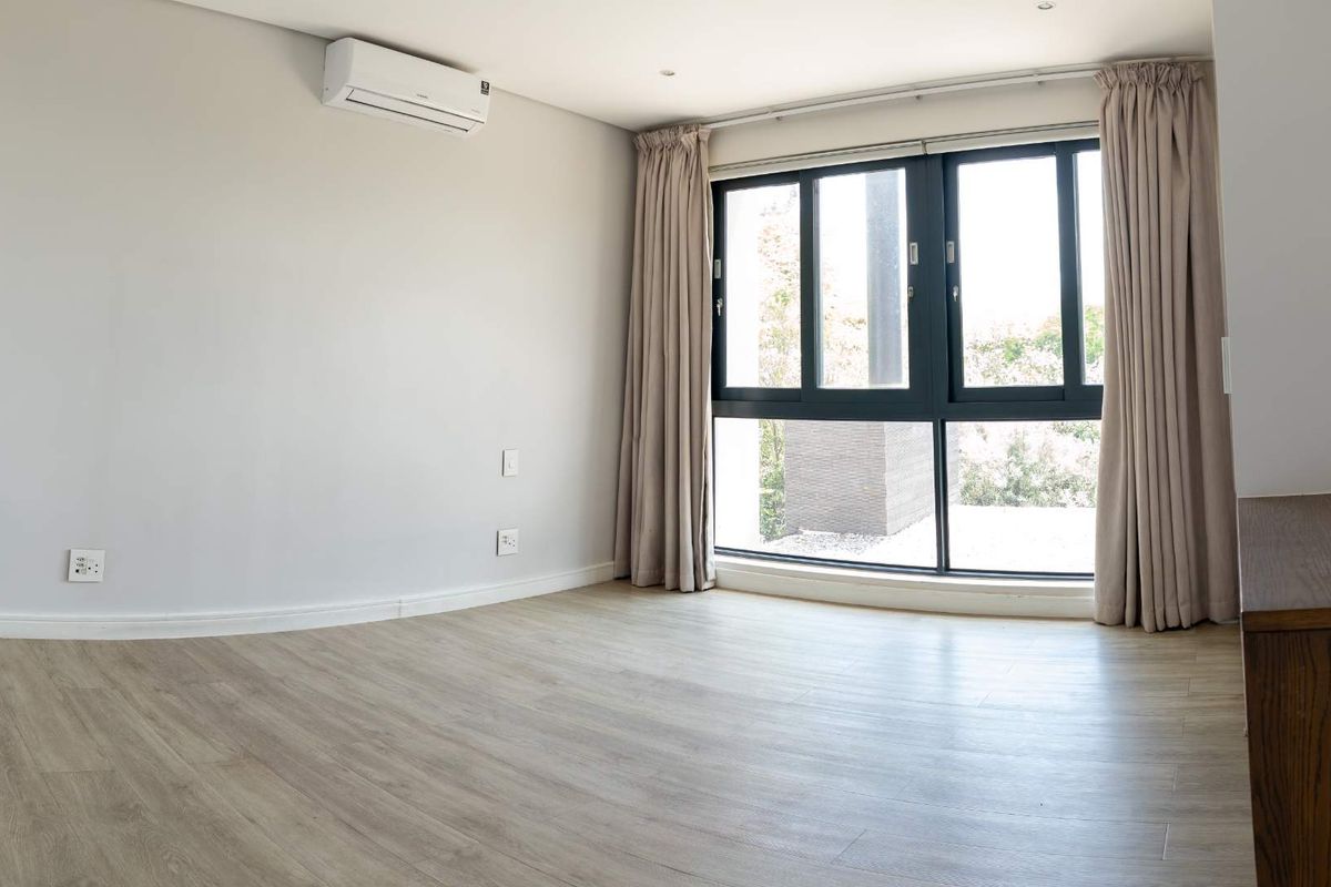 A bright, empty bedroom featuring light wood-look flooring, a wall-mounted air conditioning unit, and large windows with neutral curtains.