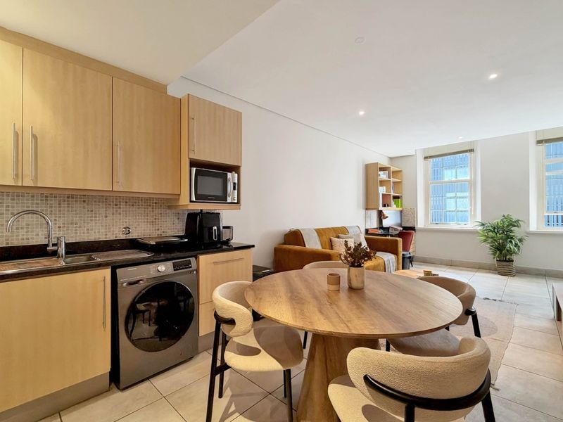 A bright open-plan living area featuring a modern kitchen with light wood cabinetry, stone countertops, and tiled flooring throughout.