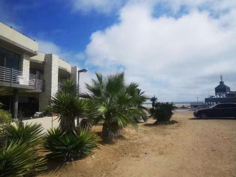Front exterior view of a modern multi-story property featuring stone cladding and balcony access, situated near the coast with an unpaved approach.