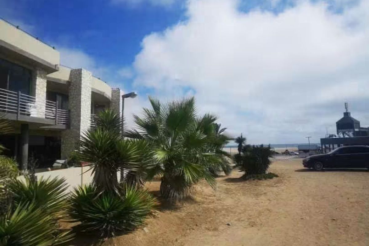 Front exterior view of a modern multi-story property featuring stone cladding and balcony access, situated near the coast with an unpaved approach.