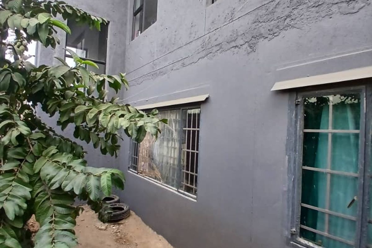 A view of the rear exterior wall featuring grey textured plaster, aluminium window frames with security bars, and an unfinished ground area.