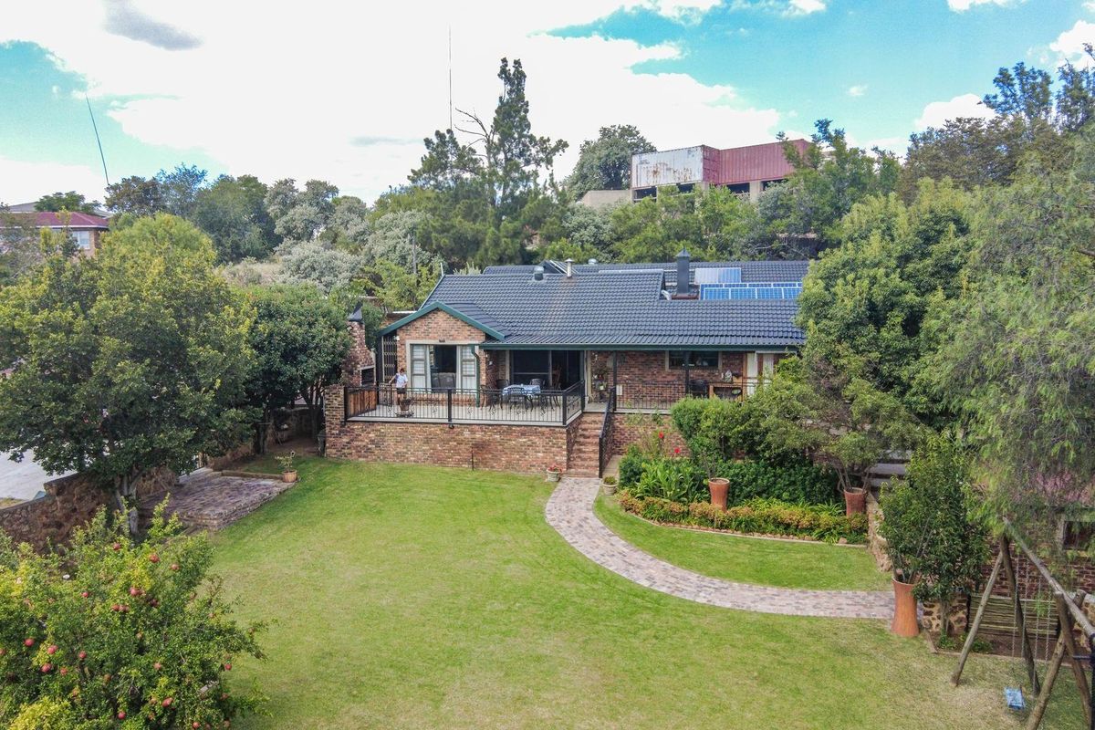 Aerial view of the rear elevation featuring a brick facade, raised patio with metal railing, tiled roof with solar panels, and a landscaped garden.