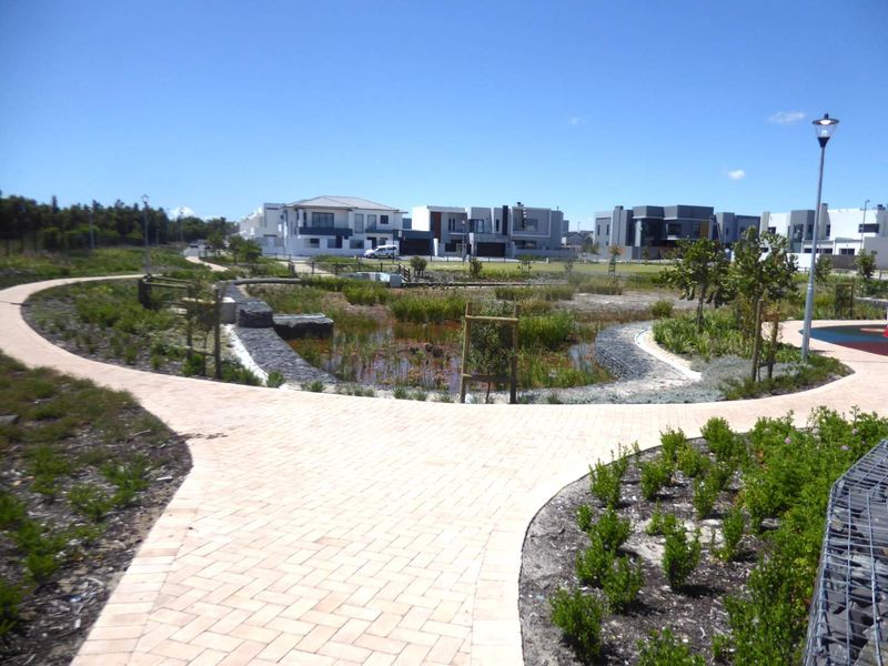 A well-maintained communal garden area featuring paved walkways, gabion retaining walls, and a landscaped pond within a residential estate.