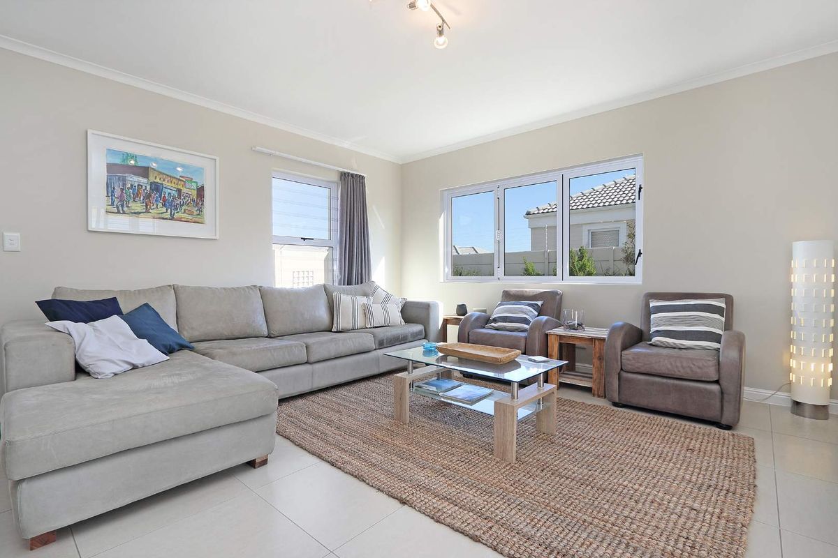 A bright and well-maintained living room featuring neutral tiled flooring, coved ceilings, and large aluminium-framed windows allowing for ample natural light.