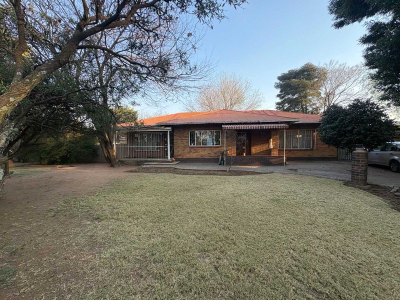 Front view of a single-story brick house featuring a corrugated metal roof, paved driveway, and established garden area.